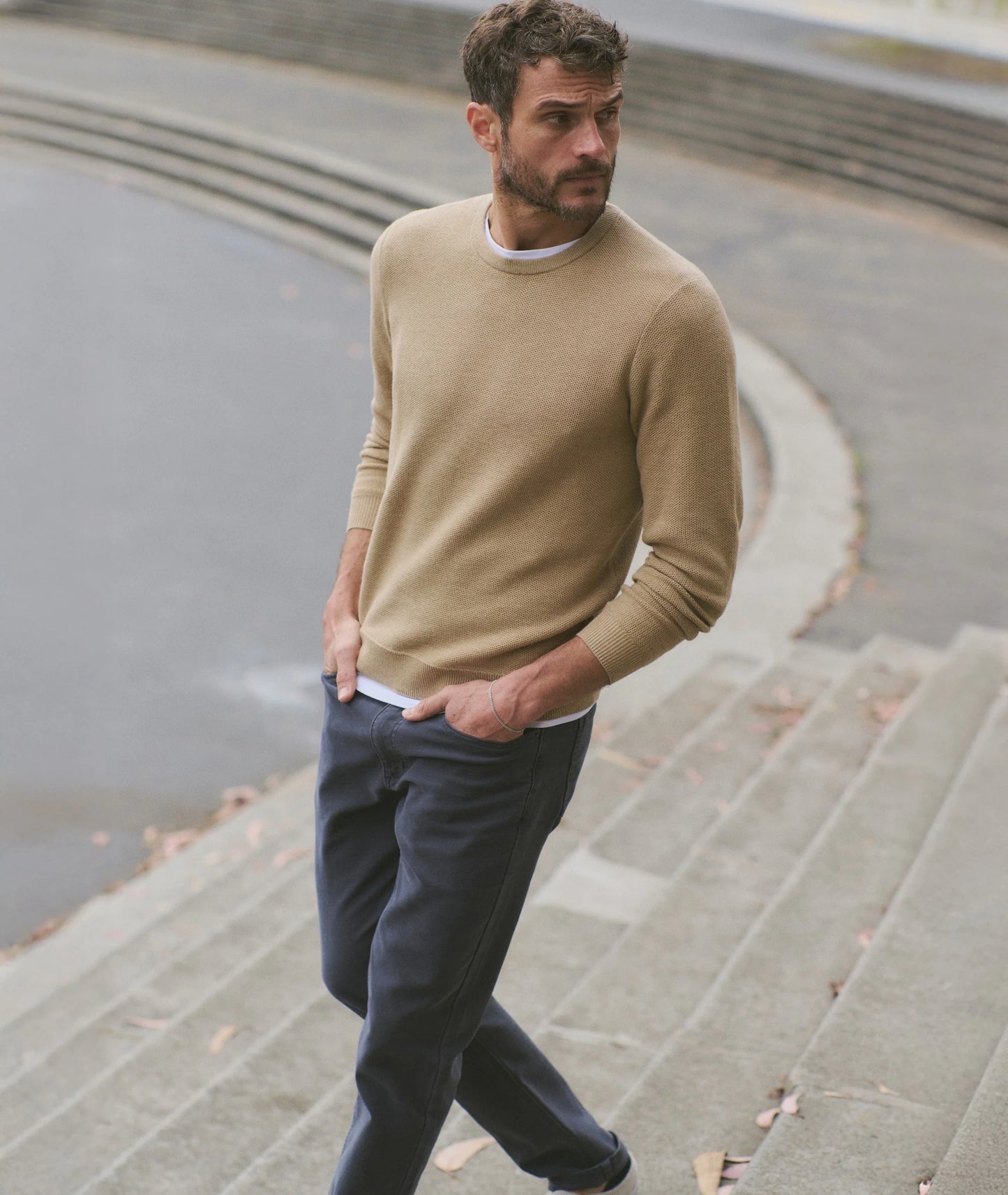 Man wearing a beige sweater and dark pants walking on steps near a body of water.