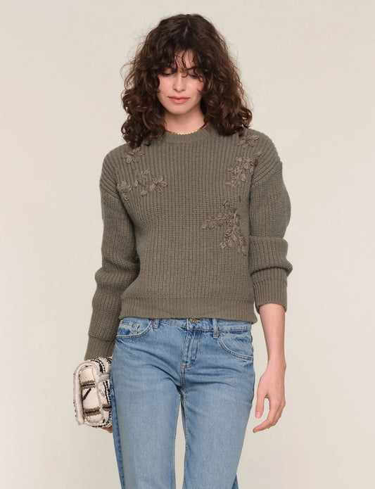 Woman wearing a green sweater with floral patterns and blue jeans on a plain background