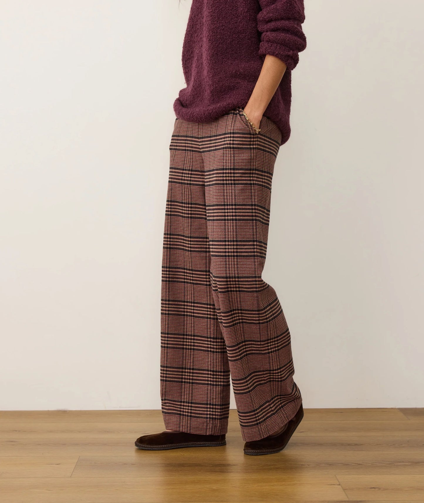Person wearing plaid pants and a maroon sweater on a wooden floor with a white wall background