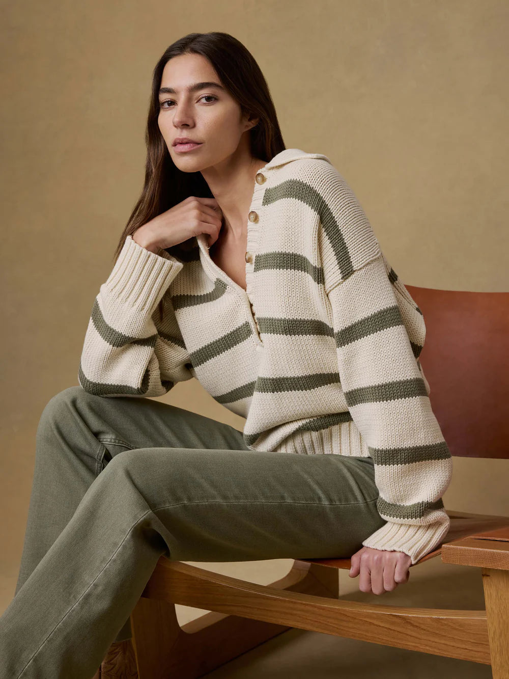 Woman wearing a striped sweater and green pants sitting on a wooden chair against a beige wall.