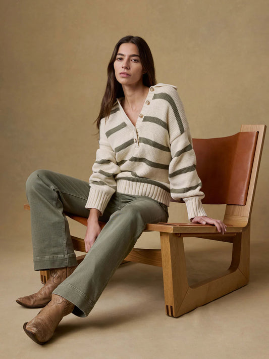 Woman sitting on a wooden chair wearing a striped sweater and green pants against a beige background