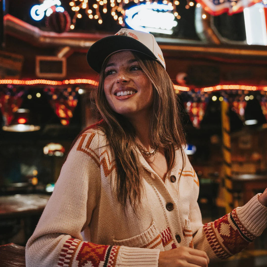 Woman wearing a patterned sweater and cap in a lively indoor setting with neon lights.