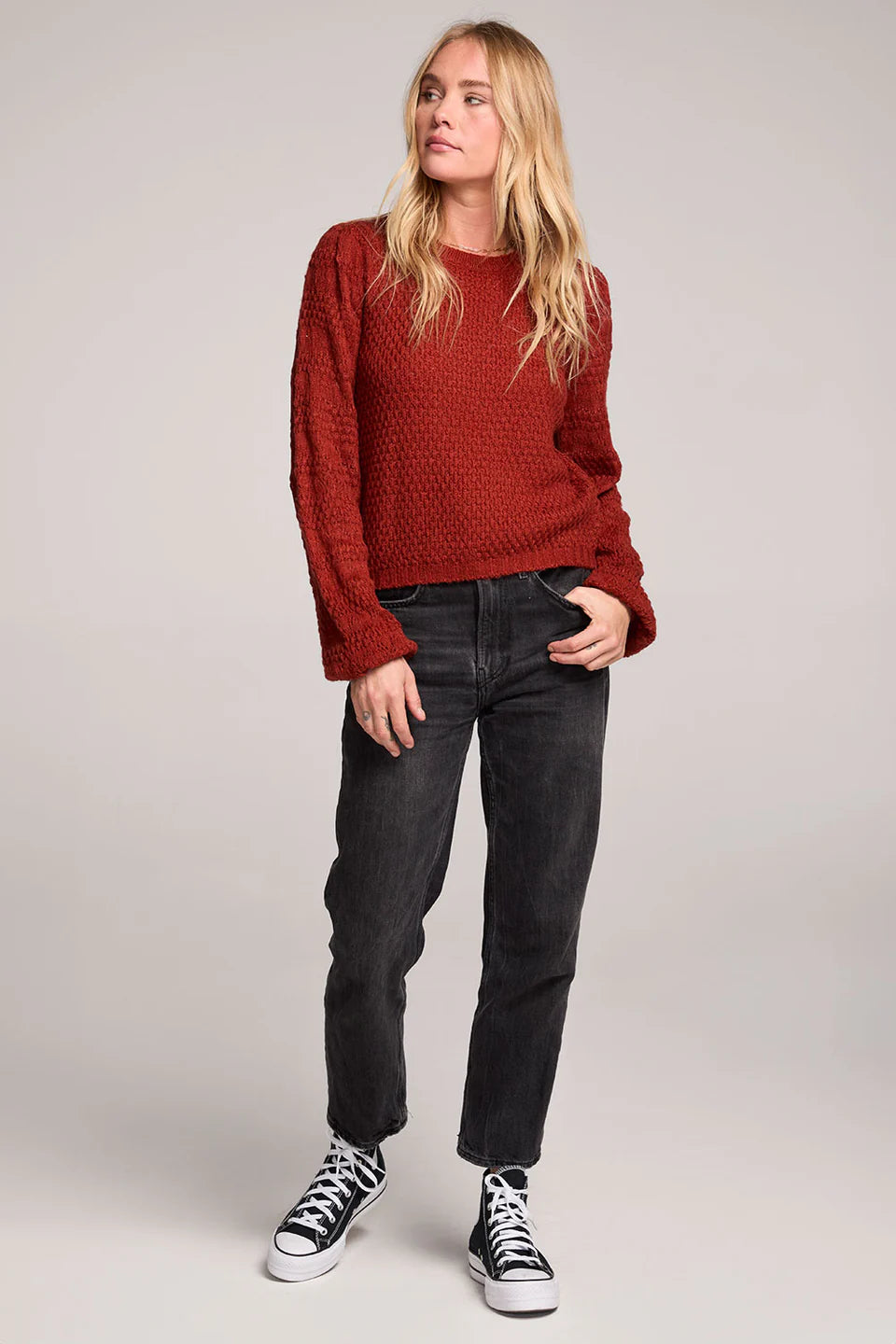 Person wearing a red textured sweater and black jeans with white sneakers on a plain background