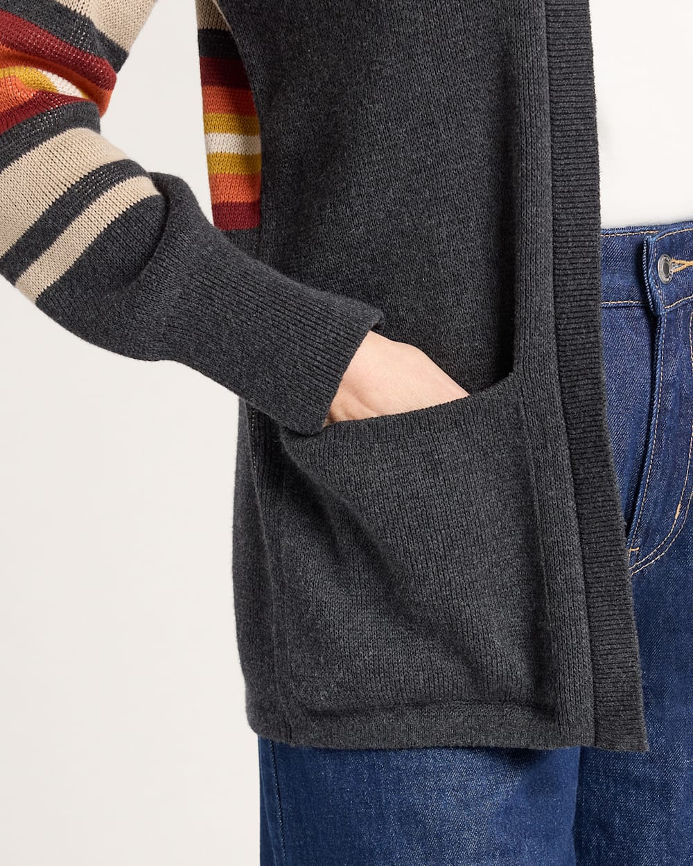 Person wearing a dark gray cardigan with a striped sweater and blue jeans on a light background