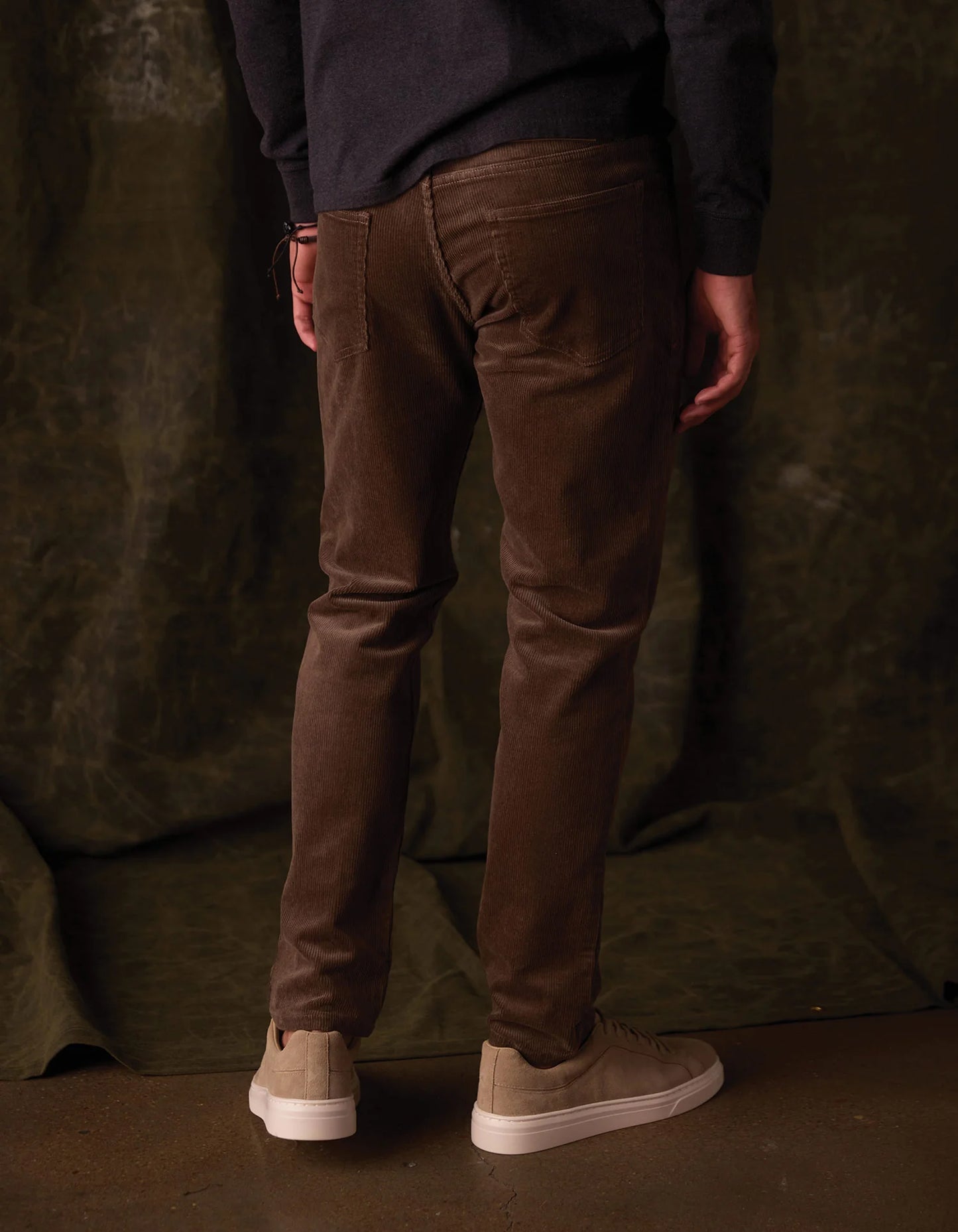 Person wearing brown pants and white shoes against a dark green curtain background