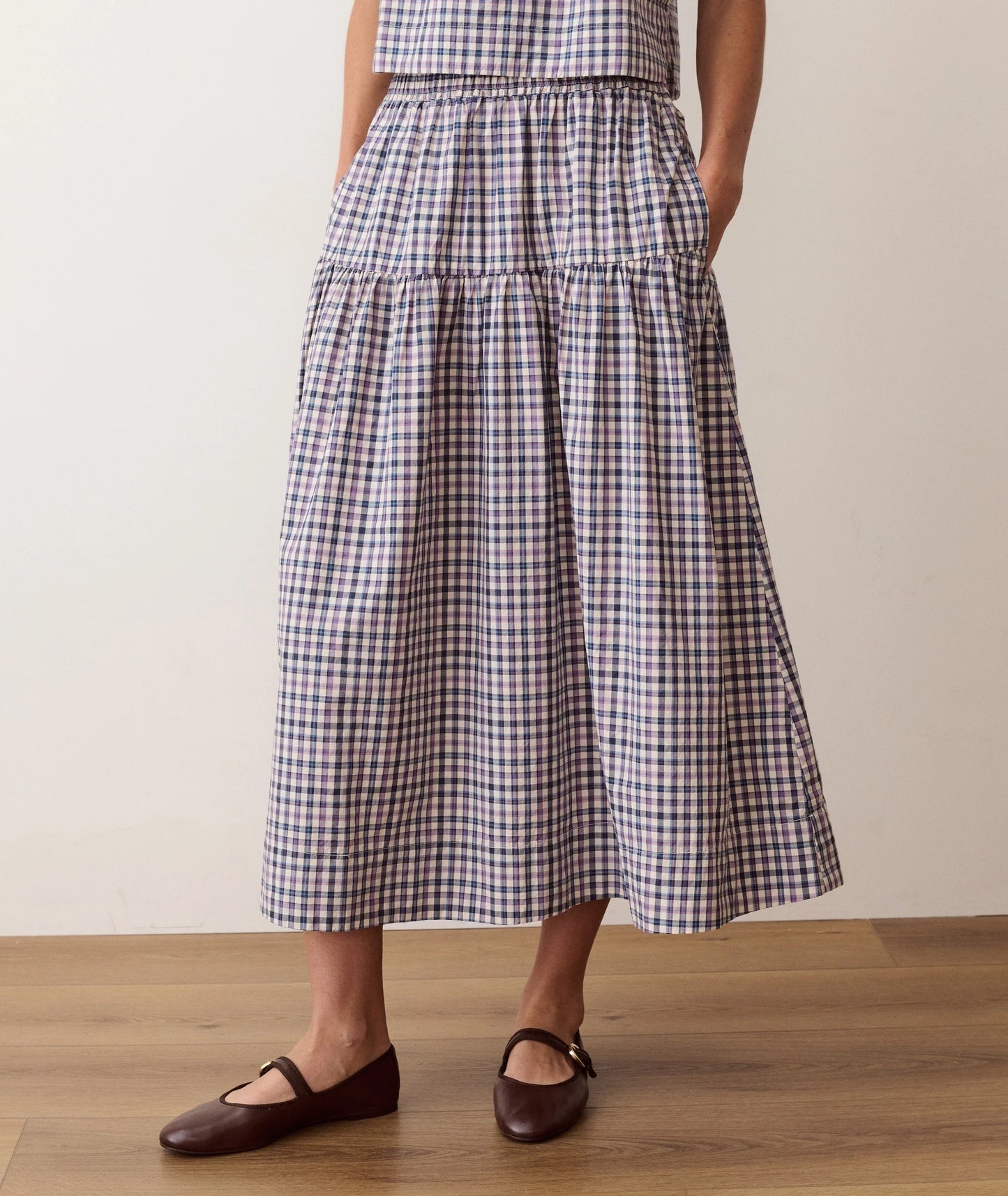 A person wearing a tiered midi skirt with a navy, beige, and white gingham check pattern, paired with dark brown Mary Jane flats, standing on a light wood floor against a plain beige wall.
