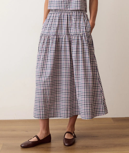 A person wearing a tiered midi skirt with a navy, beige, and white gingham check pattern, paired with dark brown Mary Jane flats, standing on a light wood floor against a plain beige wall.