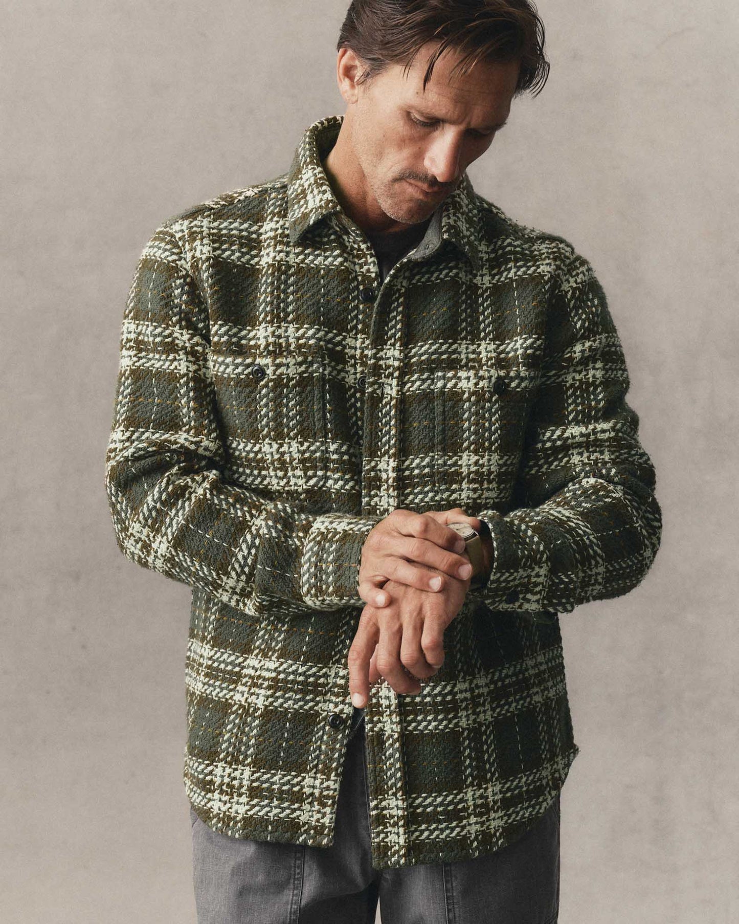 A man wearing a green and beige plaid button-up shirt jacket, looking down while standing against a textured gray wall.