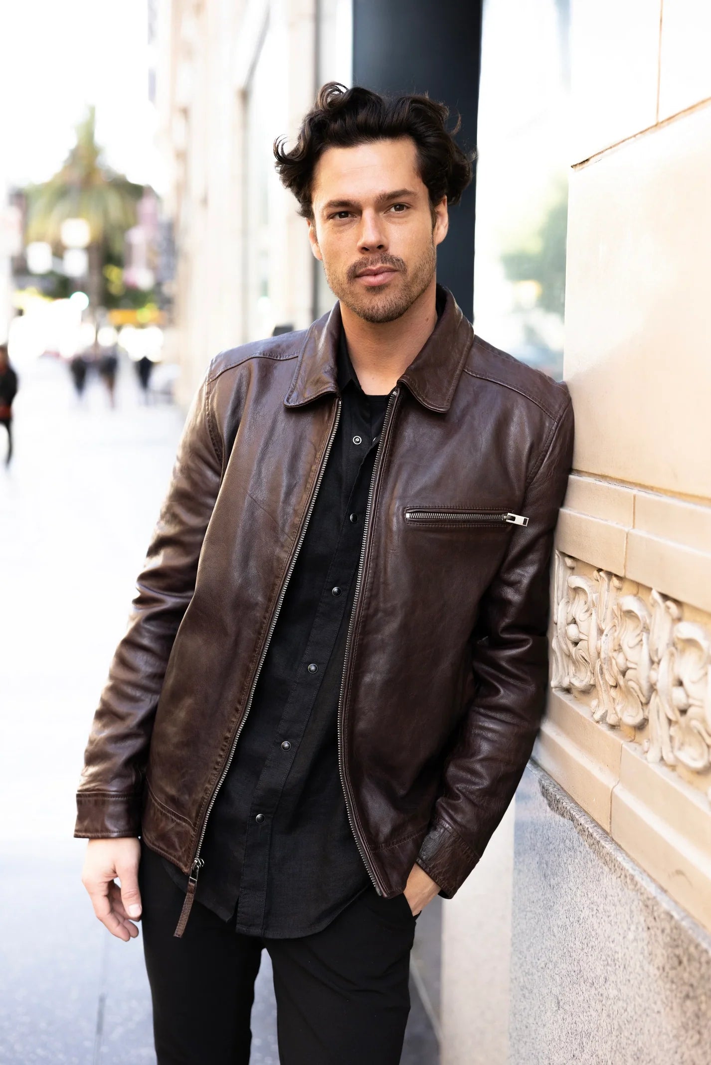 Man wearing a brown leather jacket leaning against a wall outdoors.