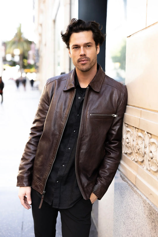 Man wearing a brown leather jacket leaning against a wall outdoors.