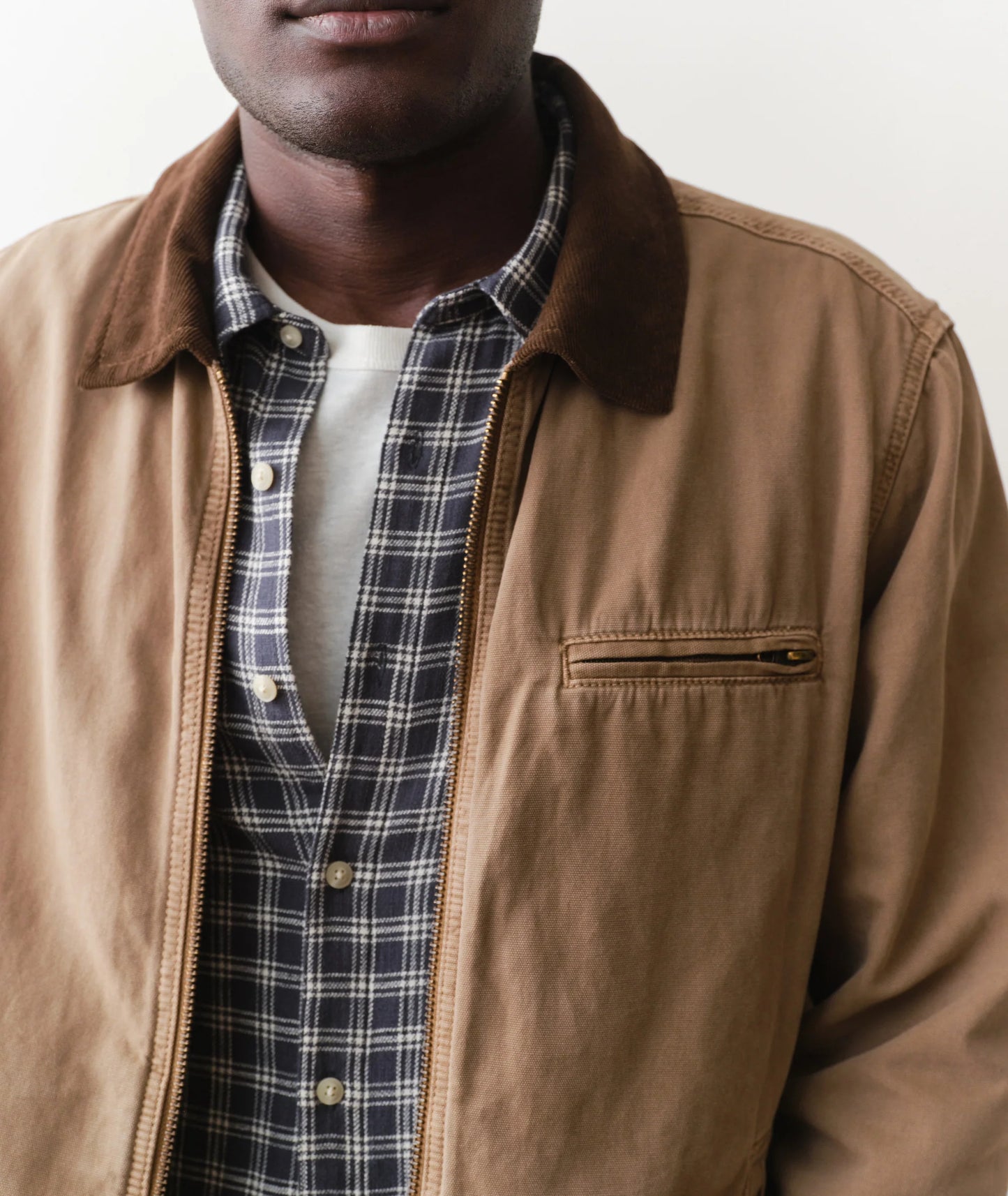 Man wearing a brown jacket over a plaid shirt with a neutral background