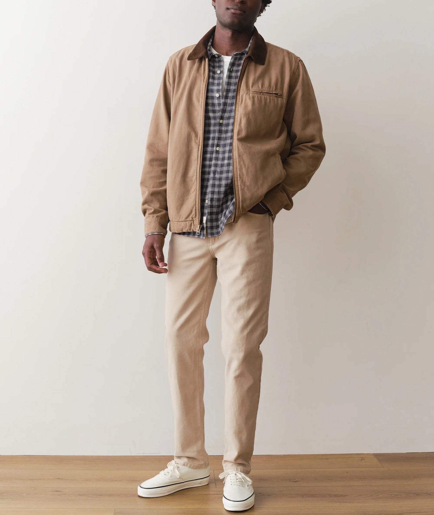 Man wearing a brown jacket, plaid shirt, beige pants, and white sneakers against a plain background