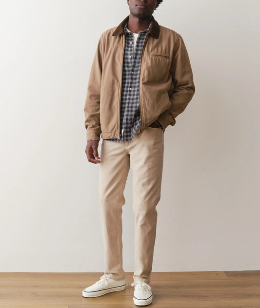 Man wearing a brown jacket, plaid shirt, beige pants, and white sneakers against a plain background