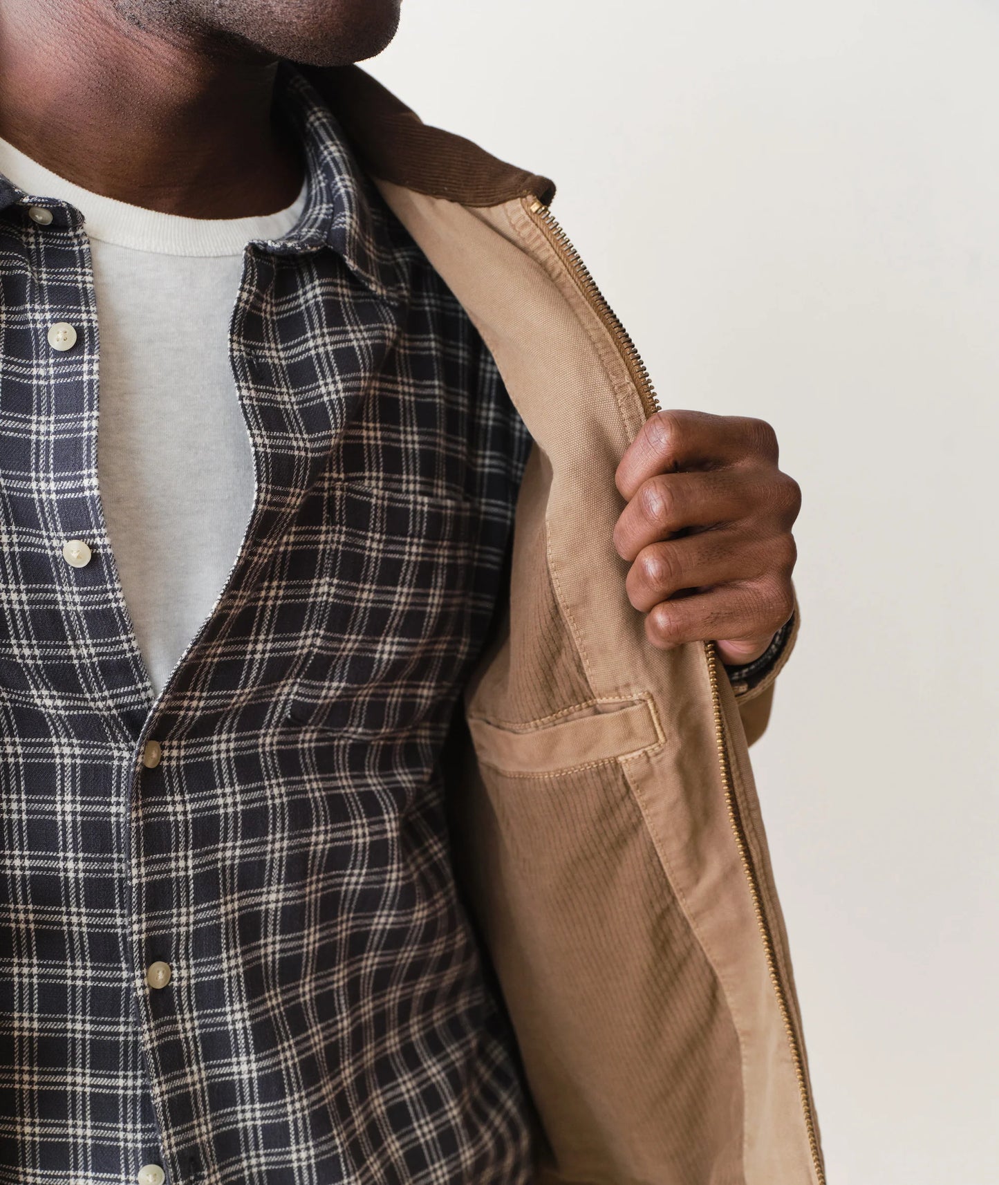 Person wearing a plaid shirt and holding a beige jacket against a neutral background