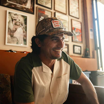 Man wearing a camouflage cap with a logo, sitting in a room with framed pictures on the wall.