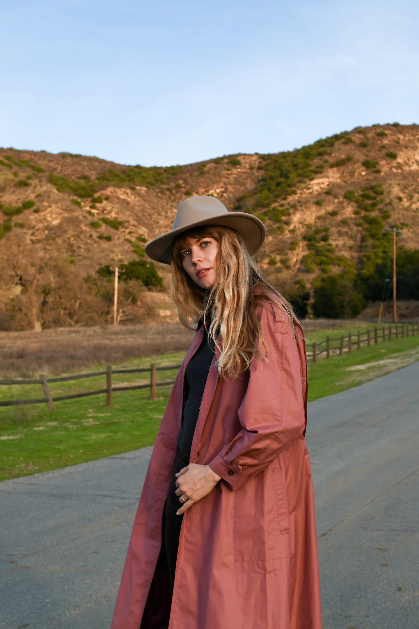 Woman in a long coat and hat standing on a road with mountains in the background