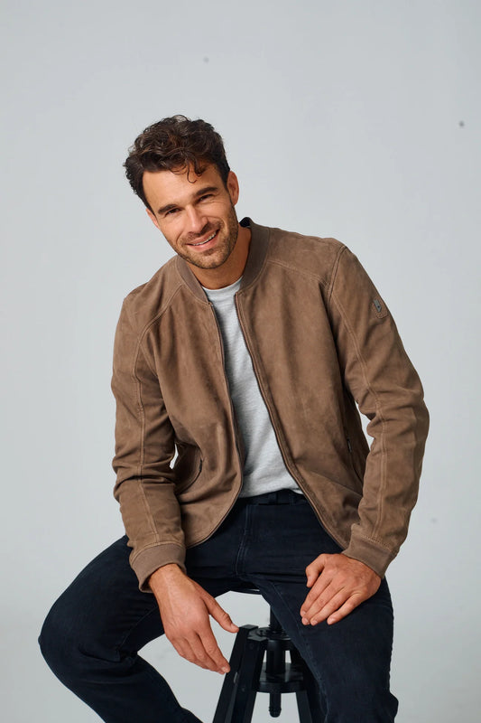 Man wearing a brown jacket sitting on a stool against a plain background