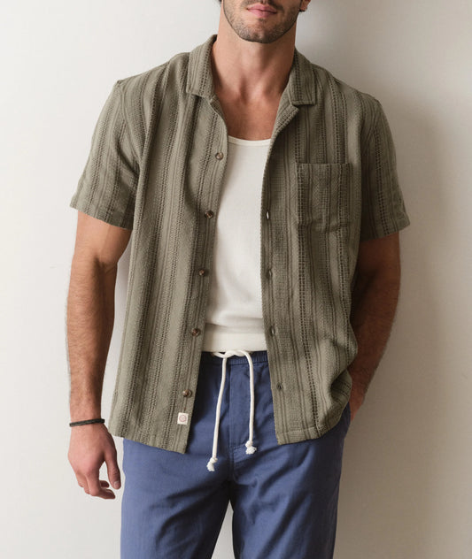 Man wearing a green patterned shirt over a white tank top and blue pants against a plain background