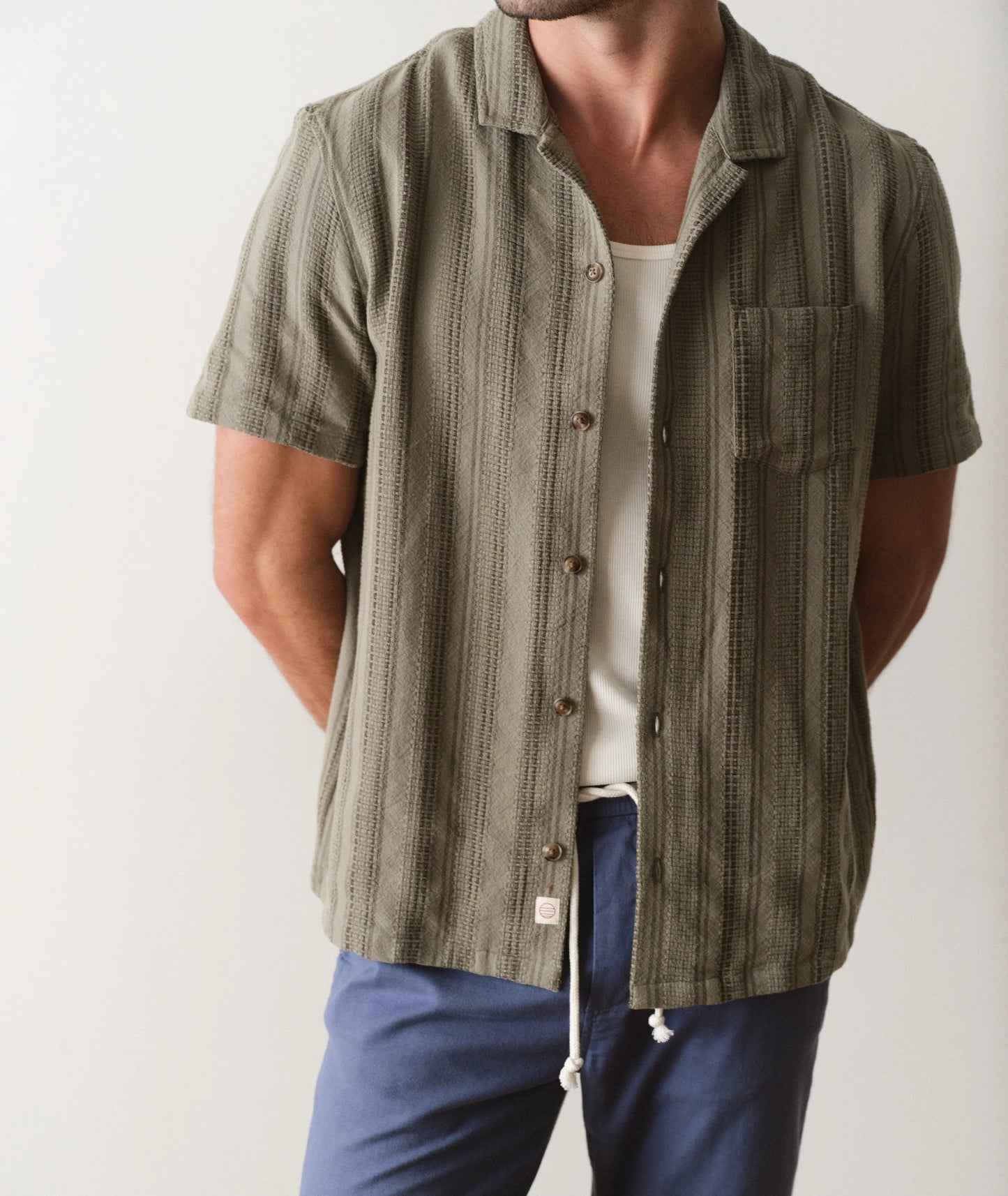 Person wearing a green textured shirt over a white tank top with blue pants on a plain background