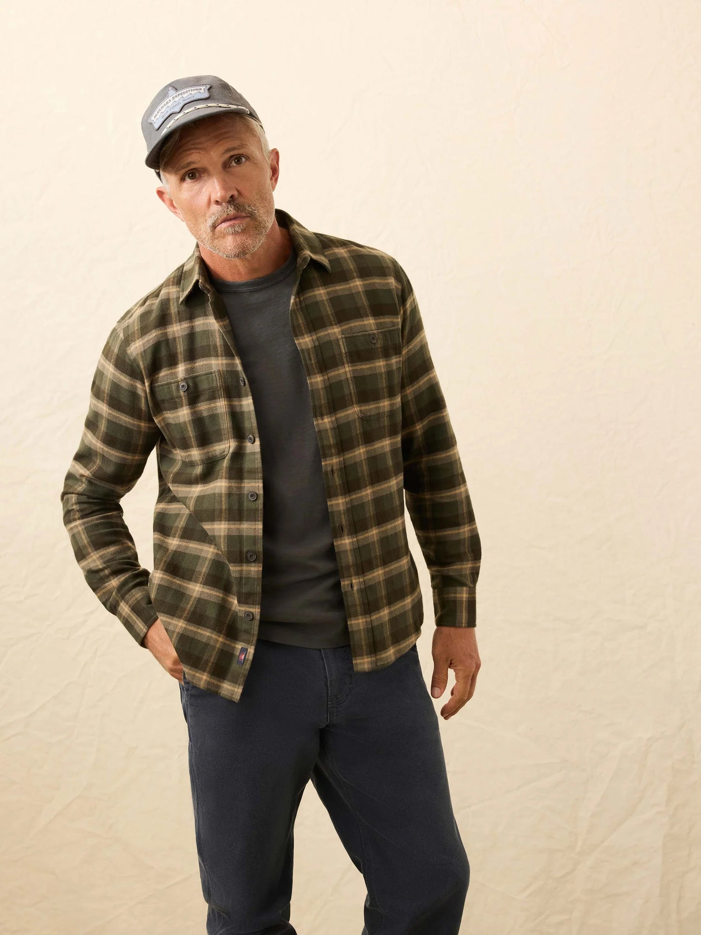Man wearing a plaid shirt, cap, and dark pants against a beige background
