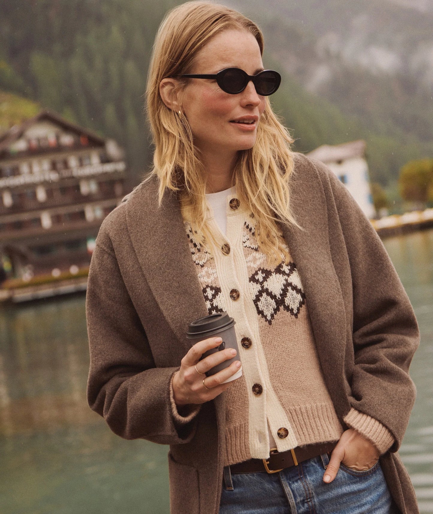 Woman in a brown coat and sunglasses holding a coffee cup, standing by a lake with mountains in the background.