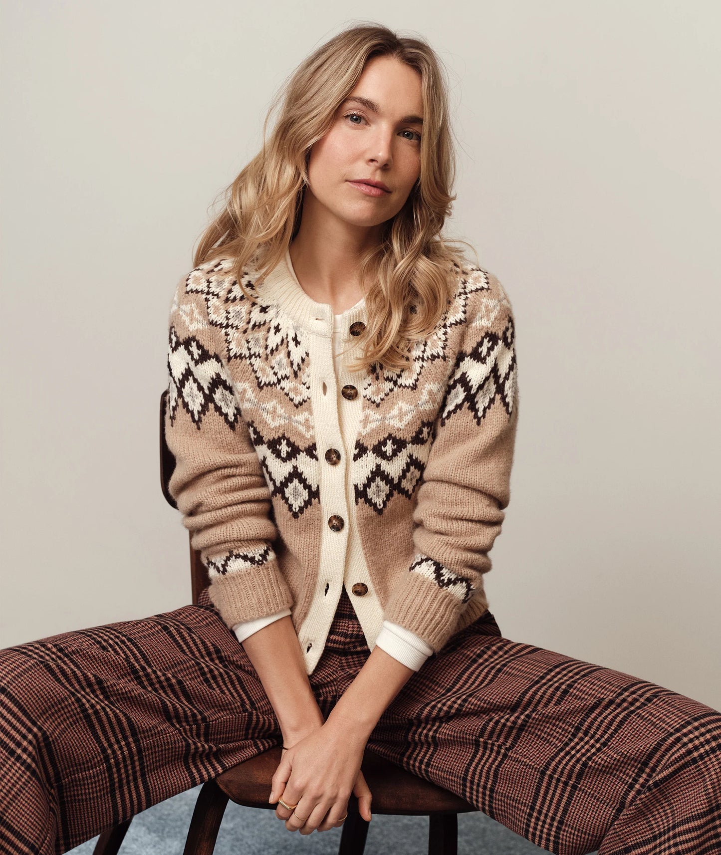 Woman wearing a patterned cardigan and plaid pants sitting on a chair against a plain background