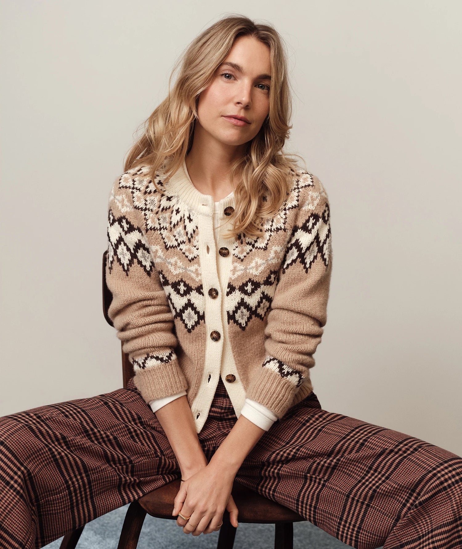 Woman wearing a patterned cardigan and plaid pants sitting on a chair against a plain background