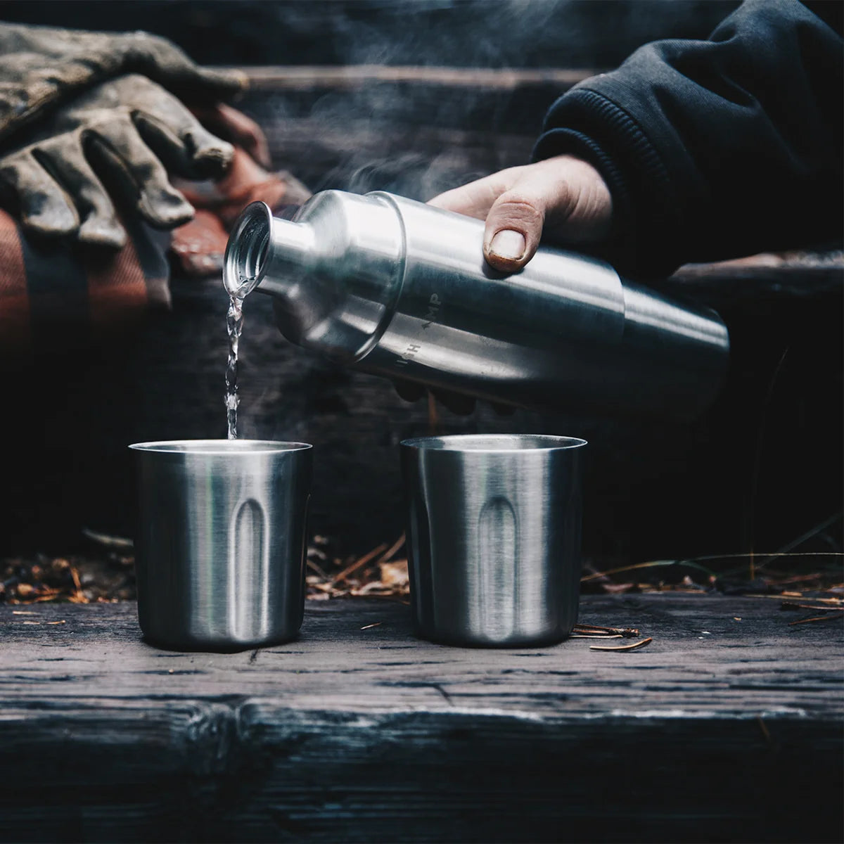 Person pouring liquid from a metal container into two metal cups on a wooden surface.