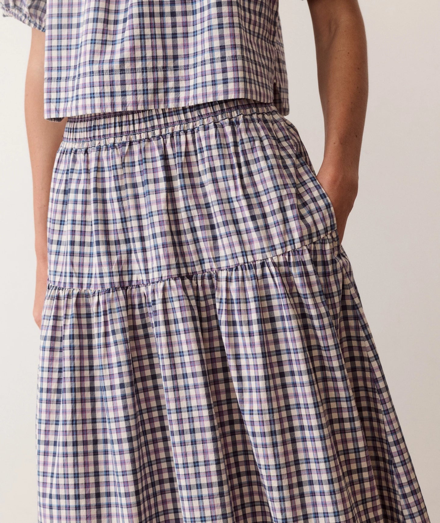 Close-up of a person wearing a tiered skirt and matching top in a blue, purple, and white gingham plaid pattern, with one hand in a pocket.