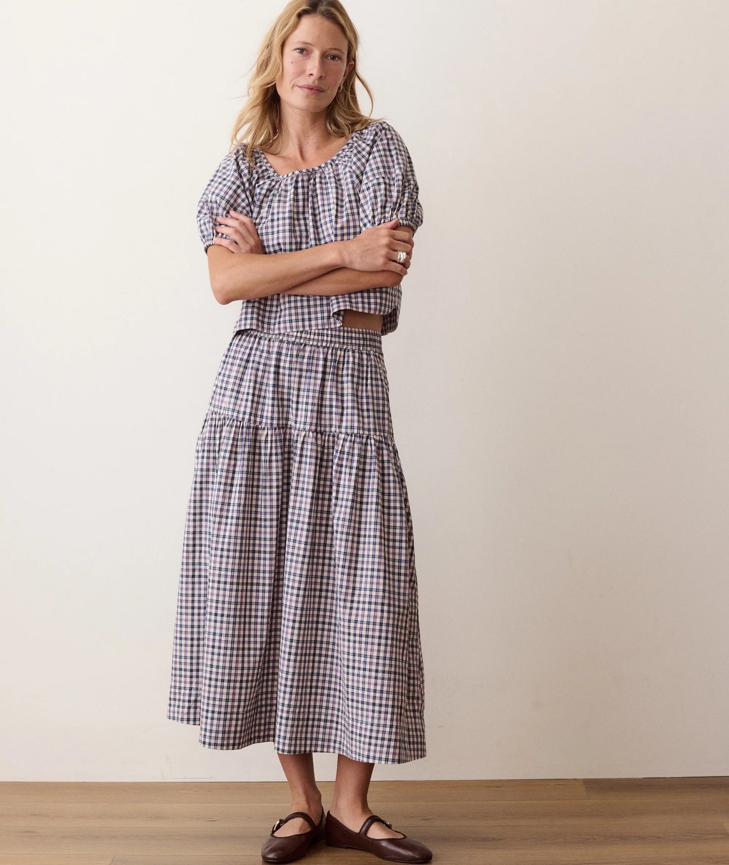 A woman modeling a matching short-sleeve puff-sleeve top and tiered midi skirt set in a black and white gingham check pattern, styled with a brown belt and dark brown flat shoes against a plain beige wall.