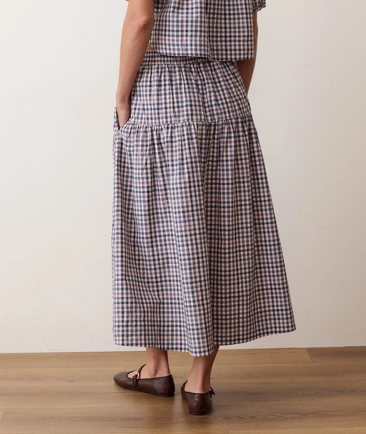 A person wearing a long tiered midi skirt with a brown, white, and black gingham check pattern, paired with dark brown flat shoes, standing on a wooden floor against a plain beige wall.