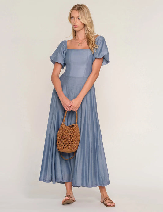 Woman wearing a blue dress holding a woven bag on a plain background