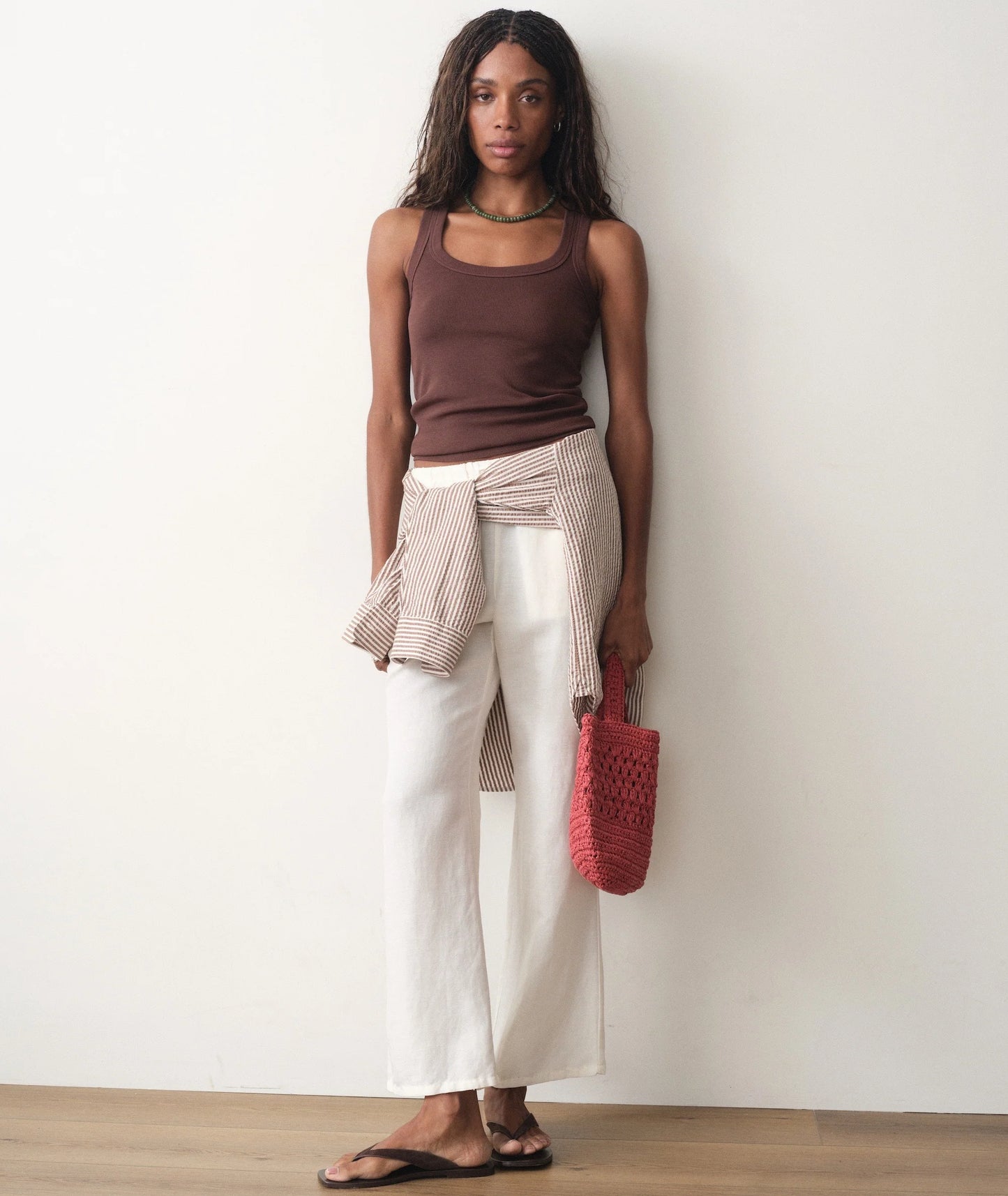 Woman wearing a brown tank top, white pants, and holding a red woven bag against a white wall.