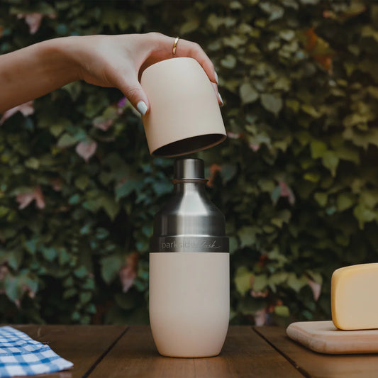 Person holding a beige cap over a silver and beige water bottle on a wooden table with a green leafy background.