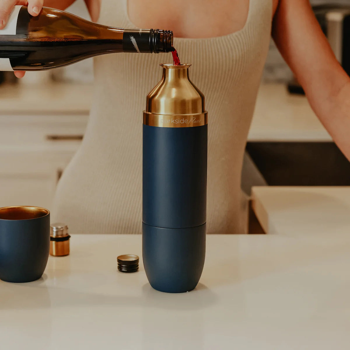 Person pouring liquid into a blue and gold cocktail shaker on a kitchen counter.
