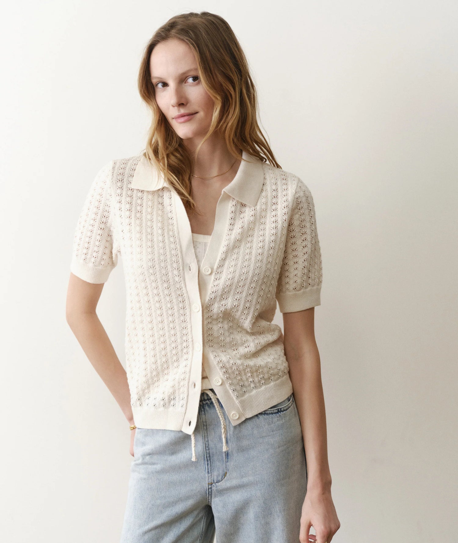 Woman wearing a light-colored knit cardigan over a white shirt with light blue jeans on a plain background
