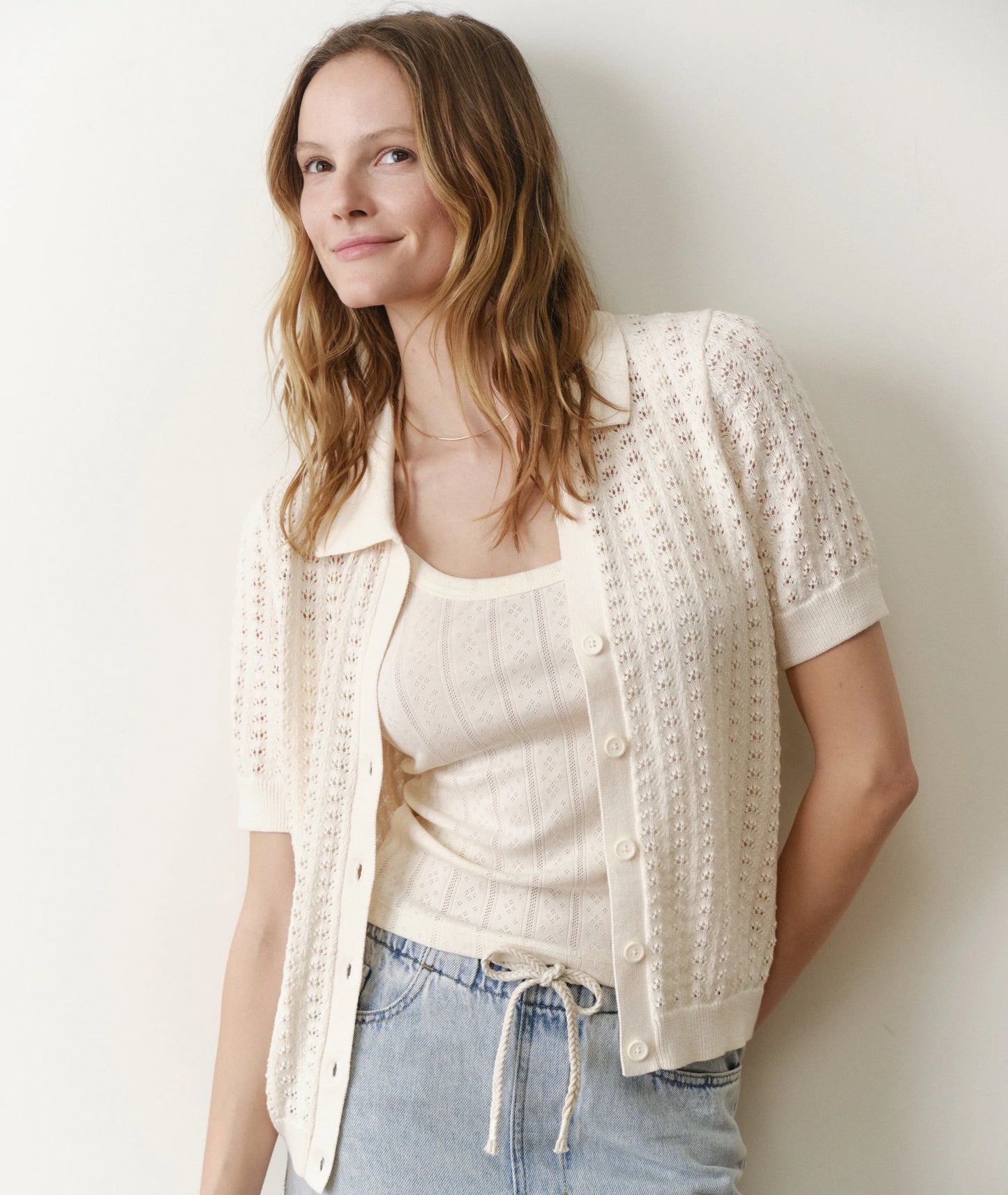 Woman wearing a cream-colored knit cardigan over a white top and blue jeans against a plain background