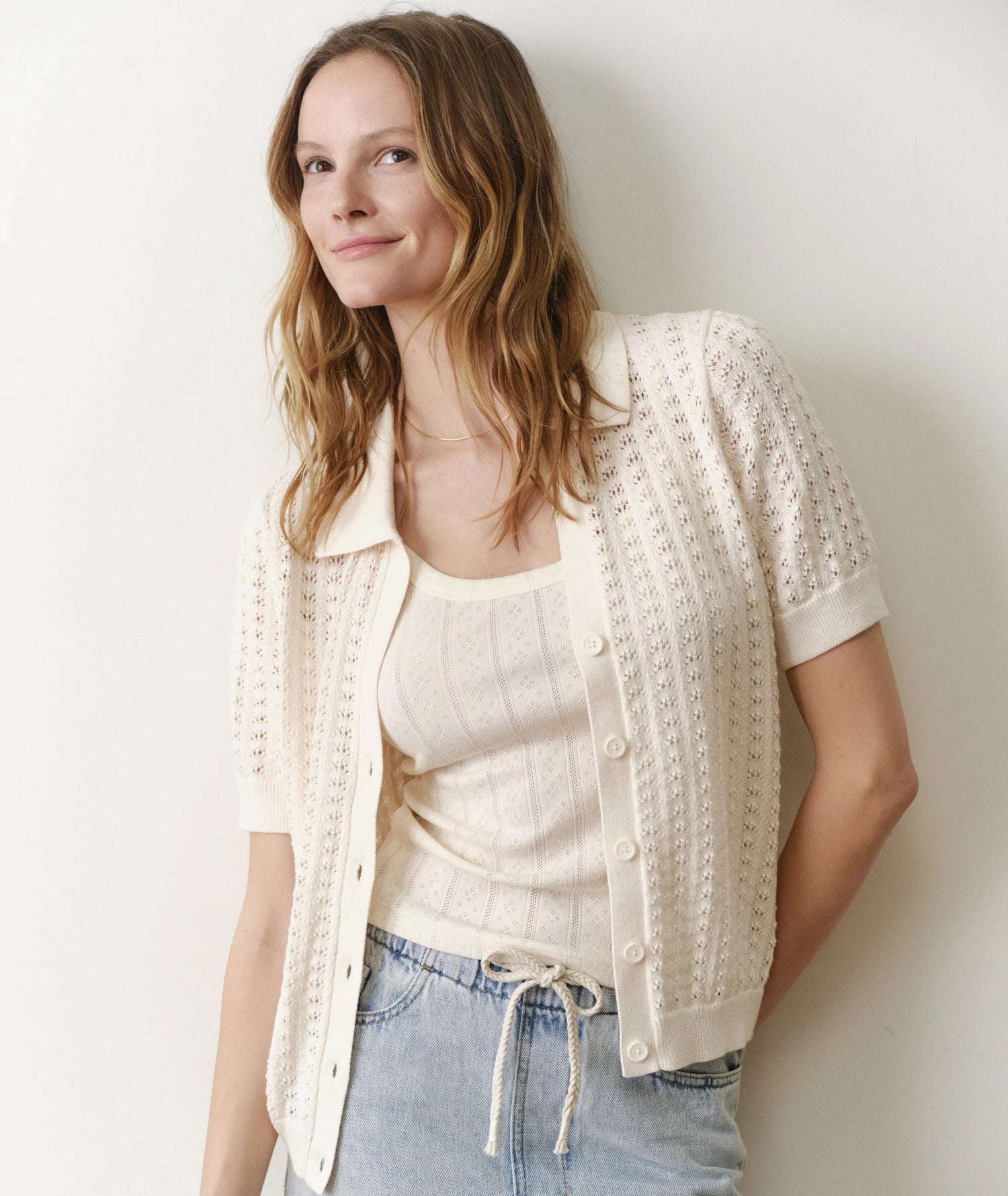 Woman wearing a cream-colored knit cardigan over a white top and blue jeans against a plain background