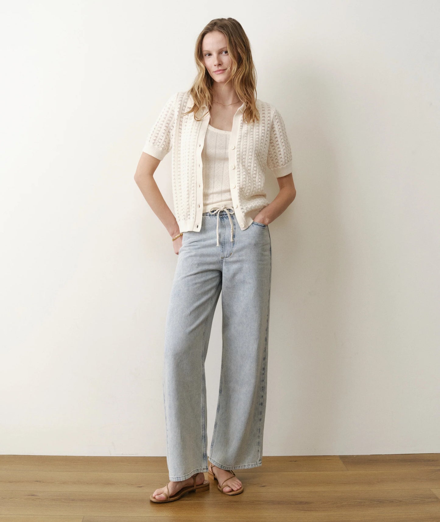 Woman wearing a white top, beige cardigan, and light blue jeans against a plain background