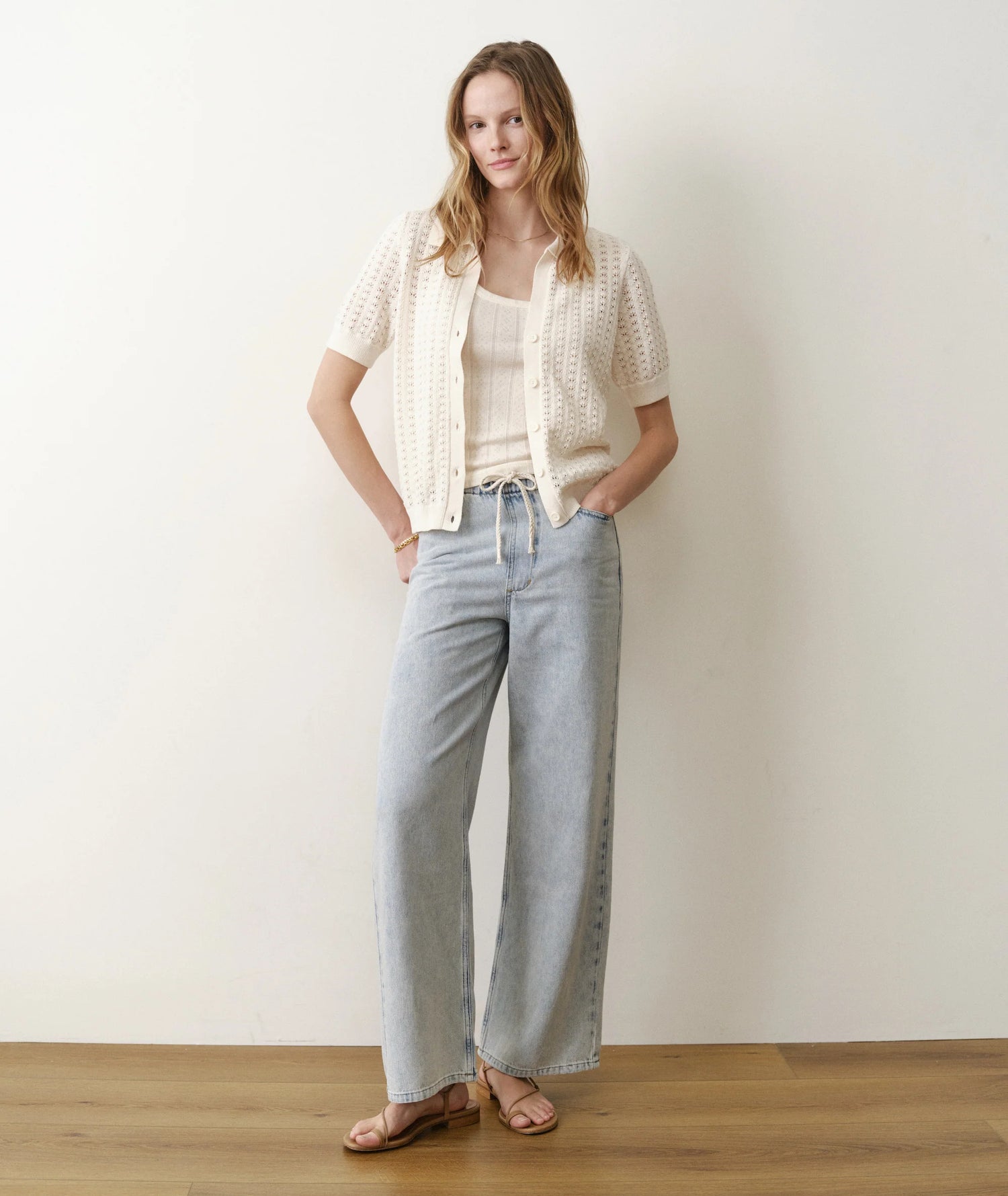Woman wearing a white top, beige cardigan, and light blue jeans against a plain background