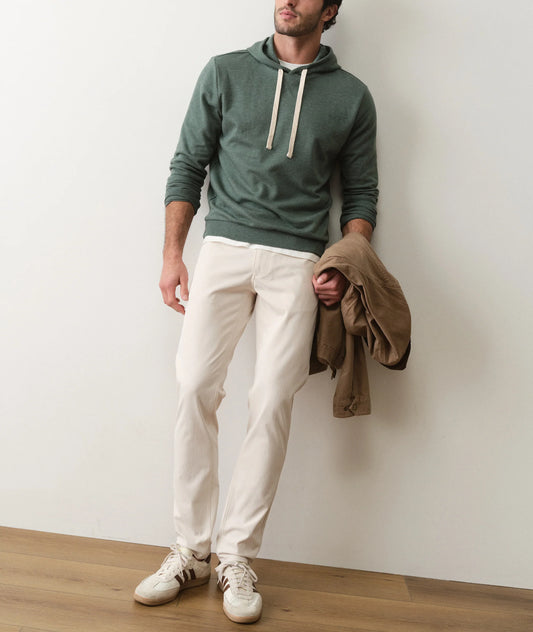 Man wearing a green hoodie, white pants, and white sneakers against a plain wall.