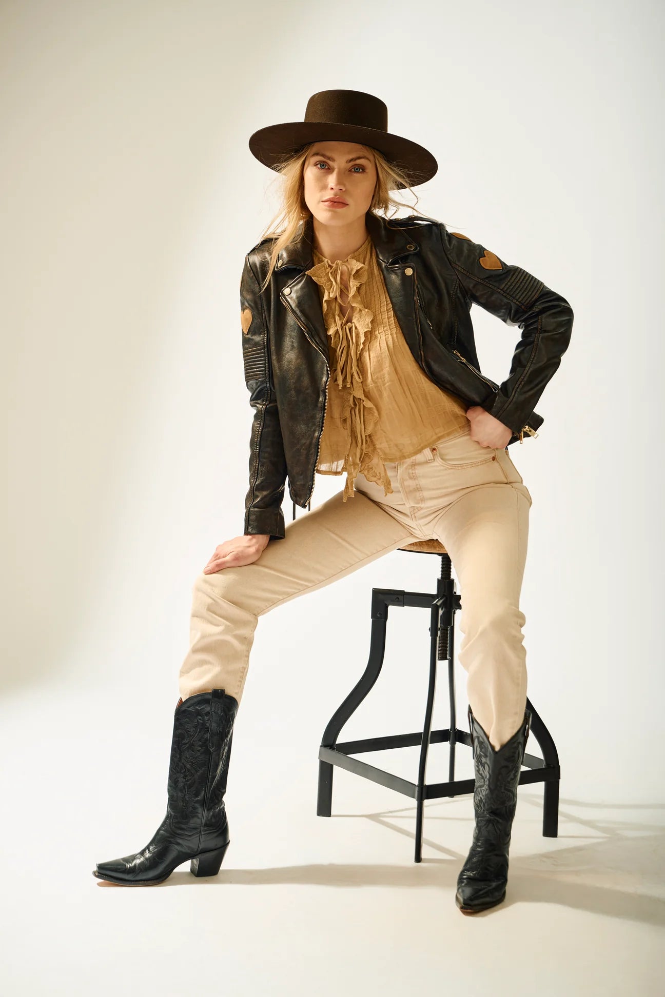 Person wearing a leather jacket, beige pants, and black boots sitting on a stool against a white background