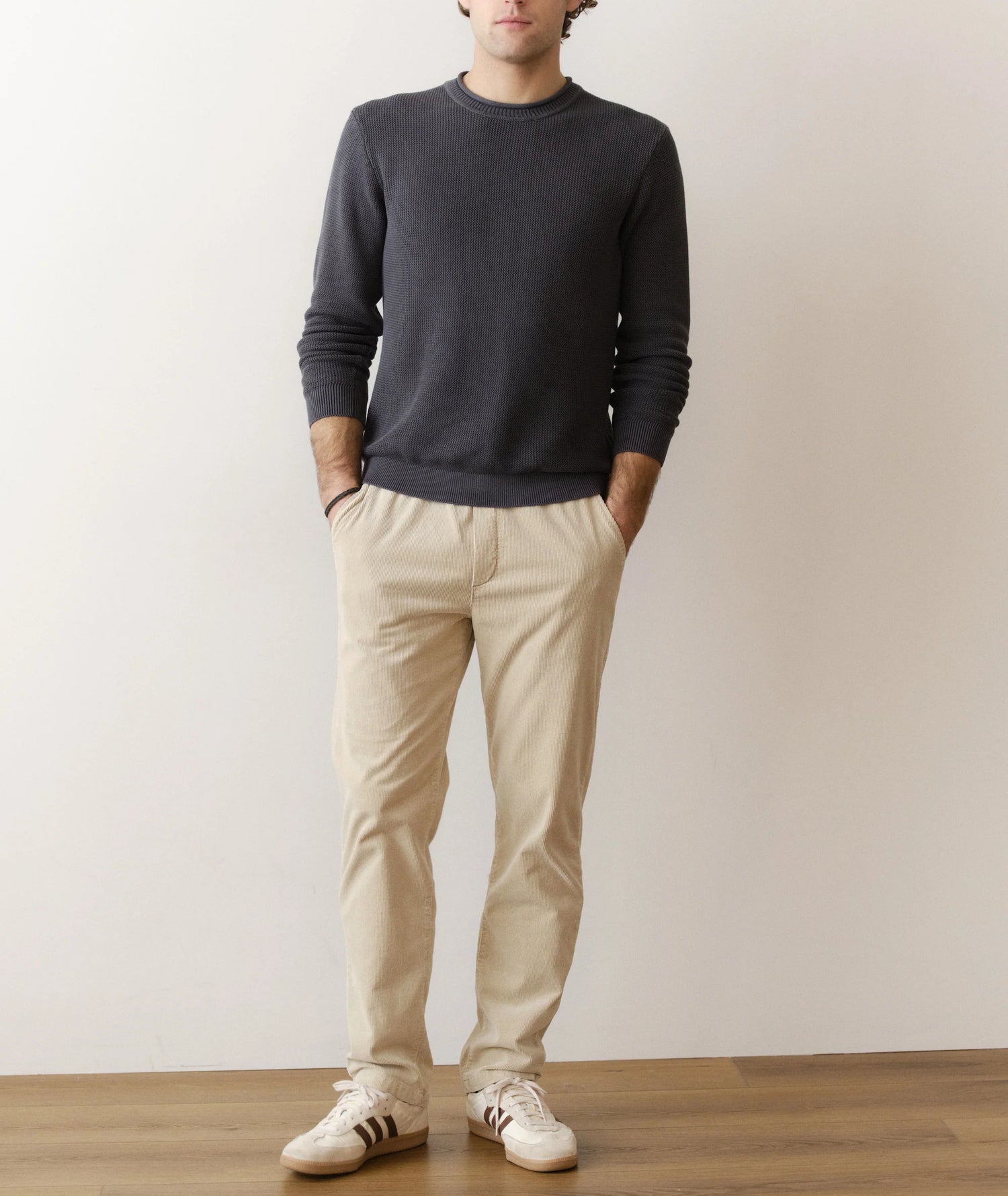Person wearing a dark gray sweater and beige pants standing against a plain white wall.