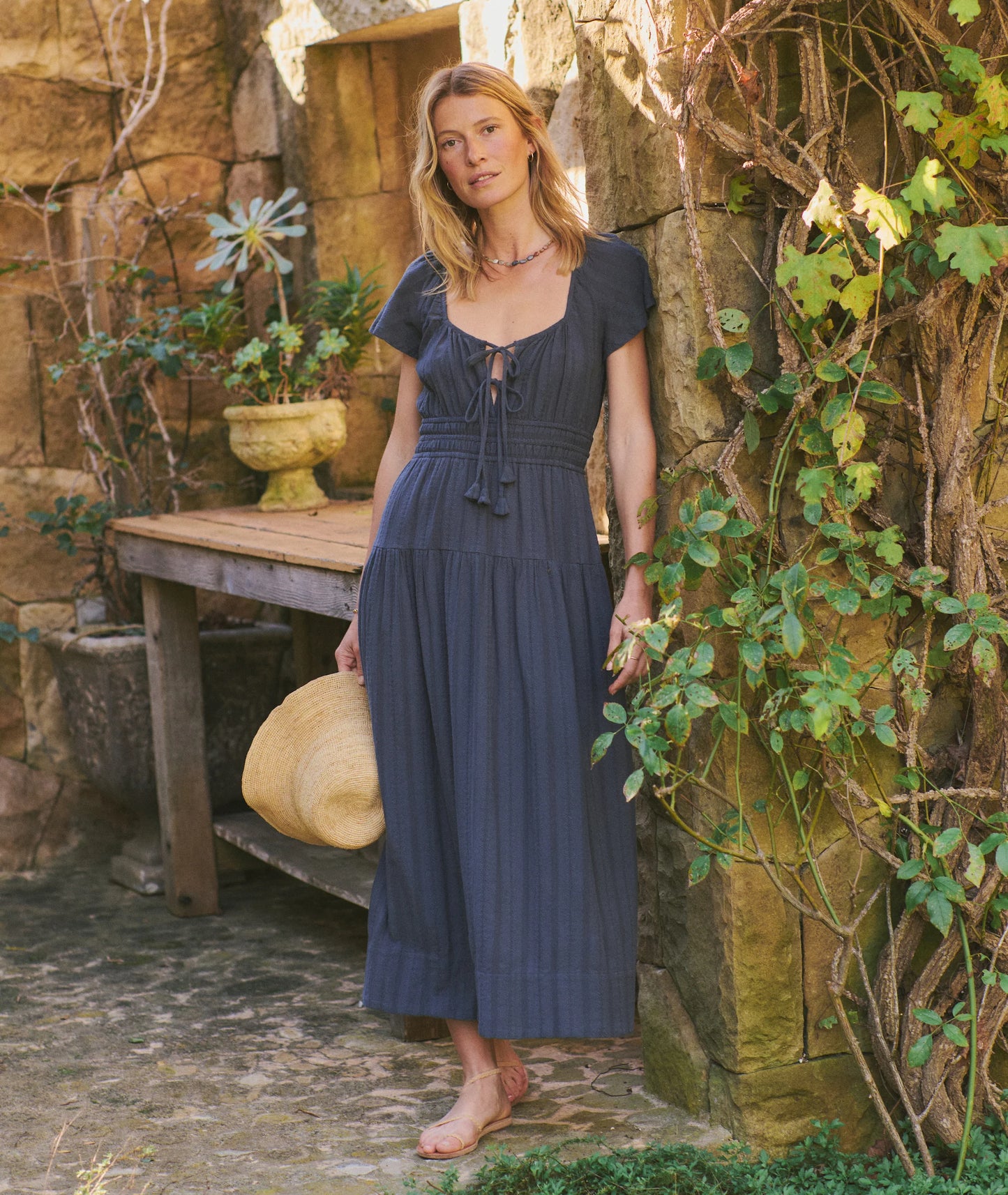 Woman in a blue dress standing in a rustic outdoor setting with stone walls and plants.