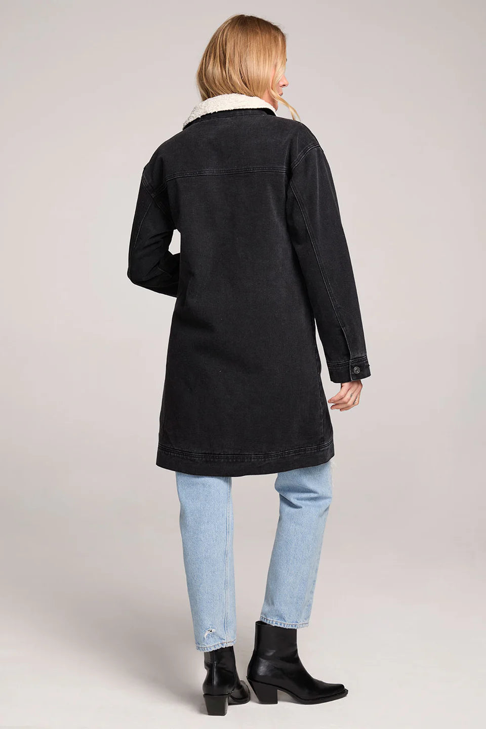 Woman wearing a black coat, light blue jeans, and black boots on a plain background