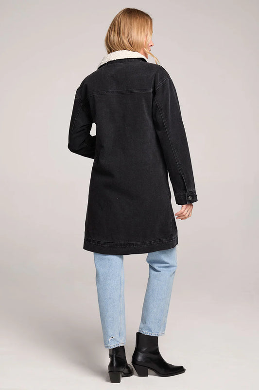 Woman wearing a black coat, light blue jeans, and black boots on a plain background