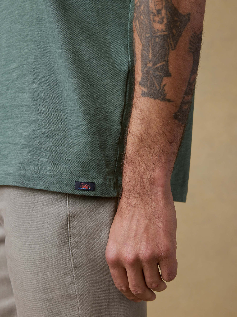 Person wearing a green shirt with a visible brand logo, beige pants, and a tattooed arm on a beige background.