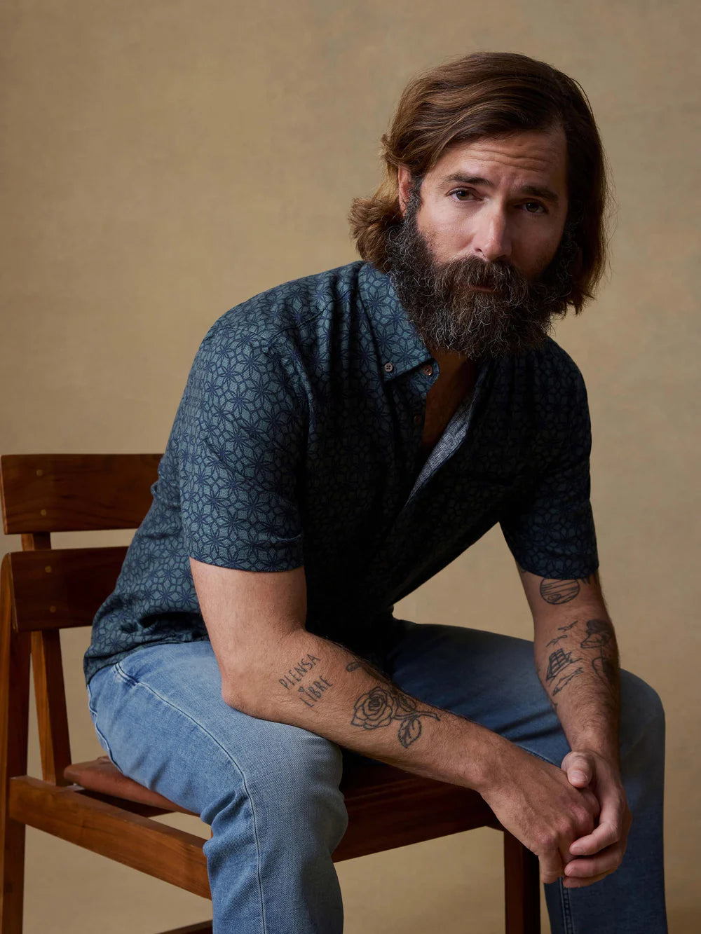 Man with a beard and tattoos sitting on a wooden chair against a beige background