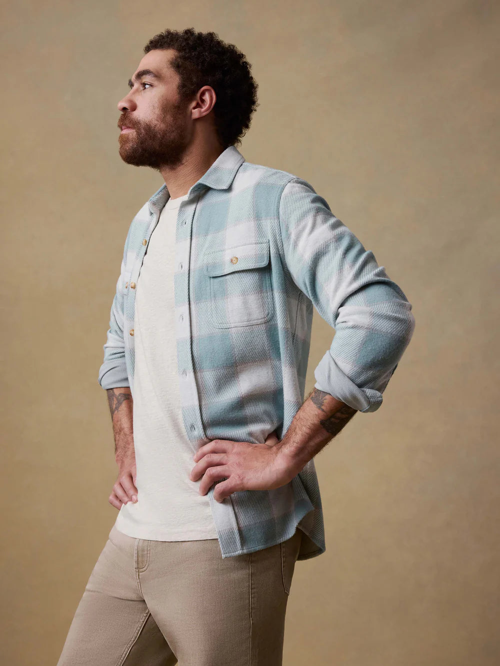 Man wearing a light blue flannel shirt over a white shirt with beige pants against a beige background