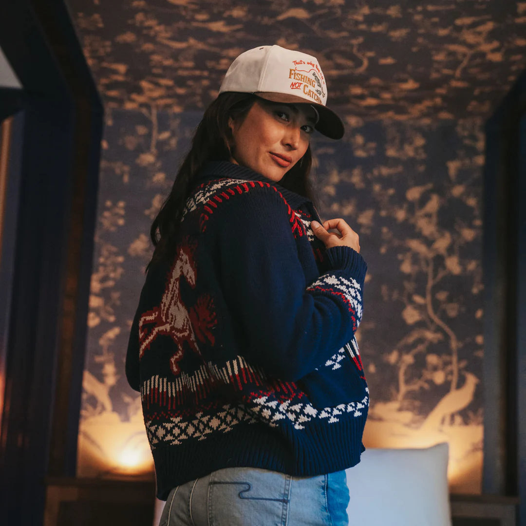 Person wearing a patterned sweater and cap in a room with decorative wall.